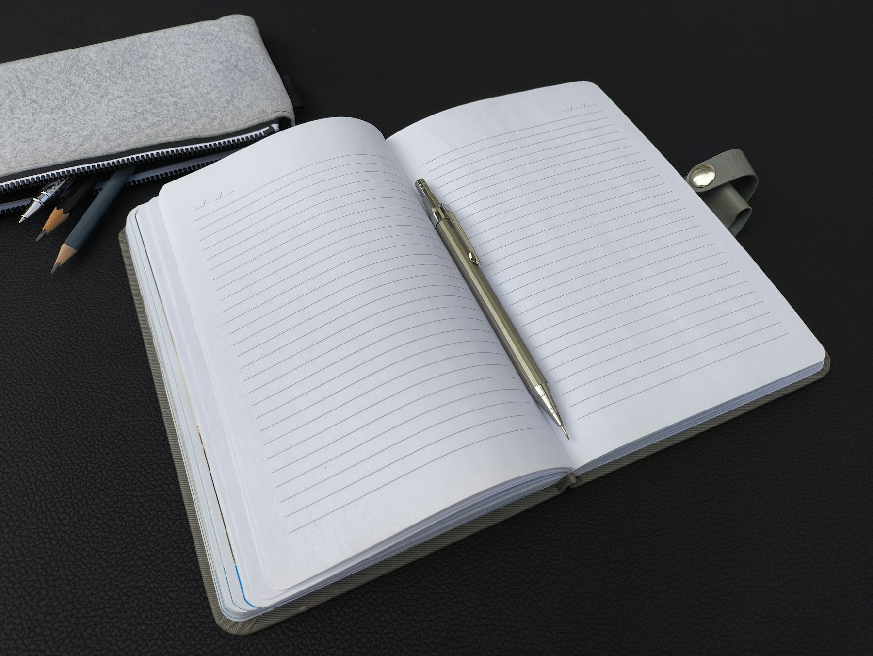 An open notebook with a pen and pencils on a dark desk.